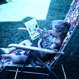 Child reading book with cat on outdoor chair.