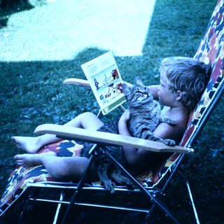 Child reading book with cat on outdoor chair.