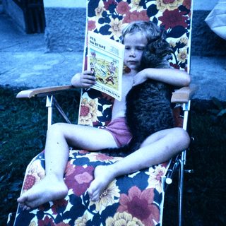 Child lounges in floral chair reading magazine.