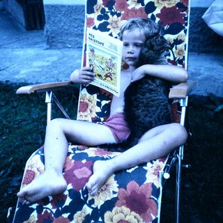 Child lounges in floral chair reading magazine.