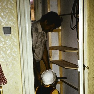 Two people carrying luggage through narrow hotel hallway.