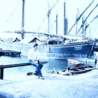 Man sitting by harbor near historic wooden boats.