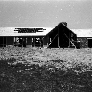 Mid-century modern house under construction on grassy hill.
