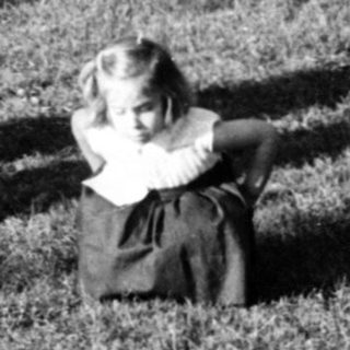 Child playing with checkers on grass, adults standing nearby in park.