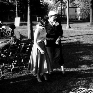 Child playing with checkers on grass, adults standing nearby in park.