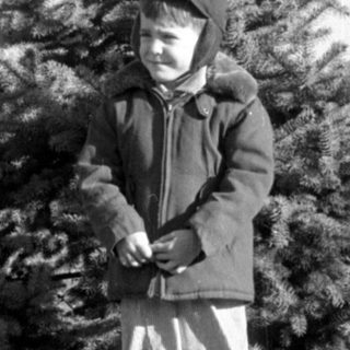 Child in winter clothing standing before an evergreen tree.