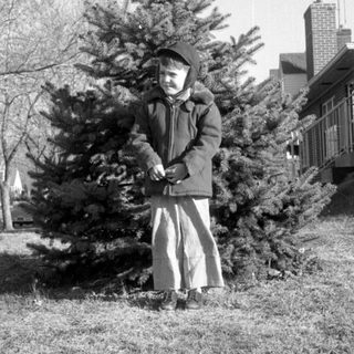 Child in winter clothing standing before an evergreen tree.