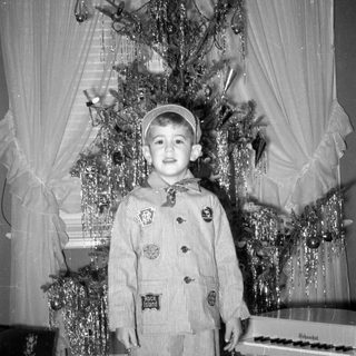 Child in striped suit standing by Christmas tree.