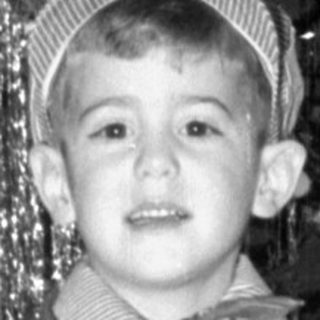 Child in striped suit standing by Christmas tree.