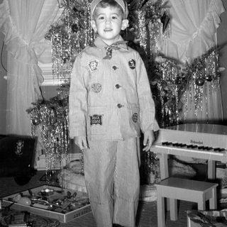 Child in striped suit standing by Christmas tree.