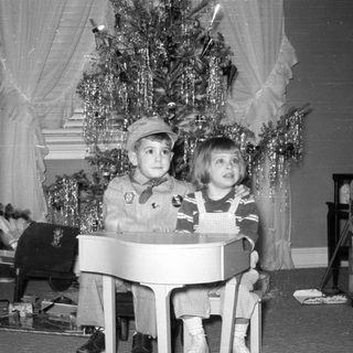 Two children with a toy piano by a Christmas tree.