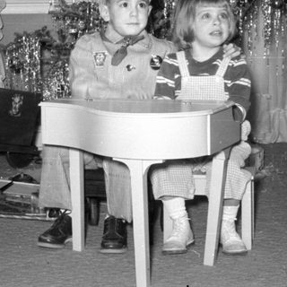 Two children with a toy piano by a Christmas tree.