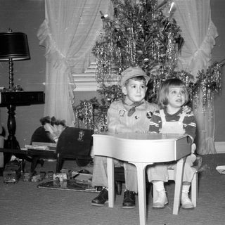 Two children with a toy piano by a Christmas tree.