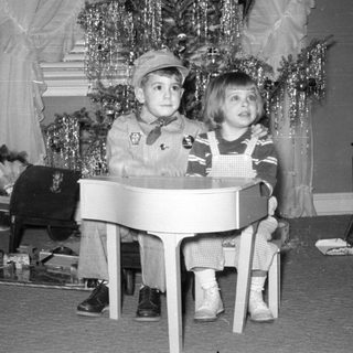 Two children with a toy piano by a Christmas tree.