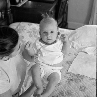 Infant on a bed with a woman and desk.