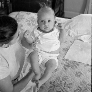 Infant on a bed with a woman and desk.