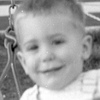 Young child in overalls sitting on a swing.
