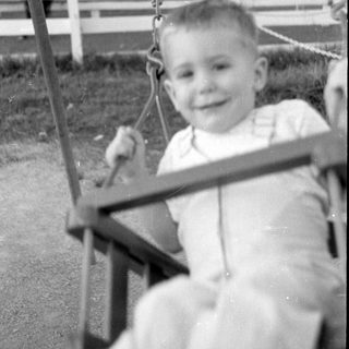 Young child in overalls sitting on a swing.