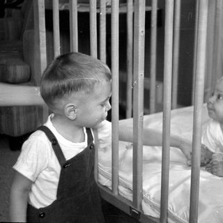 Young child in overalls standing by an infant in a crib.