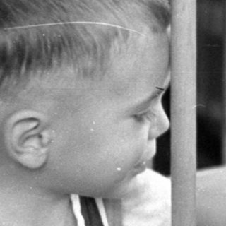 Young child in overalls standing by an infant in a crib.