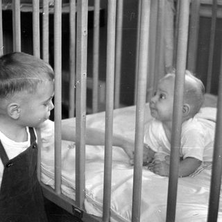 Young child in overalls standing by an infant in a crib.