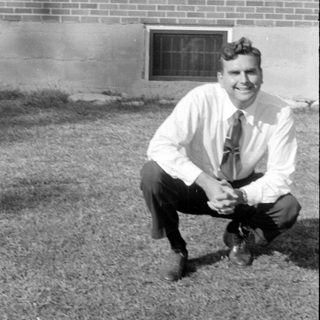 Man in dress shirt squatting in front of brick building.