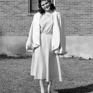 Woman in a dress and coat before a brick house.