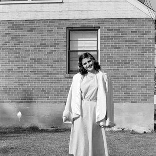Woman in a dress and coat before a brick house.