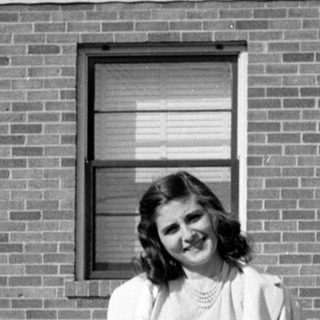 Woman in a dress and coat before a brick house.