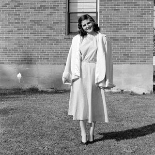 Woman in a dress and coat before a brick house.
