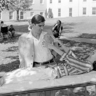 Man standing beside a woman lying on a cot.
