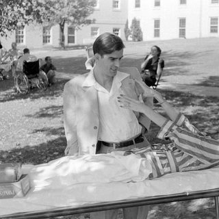 Man standing beside a woman lying on a cot.