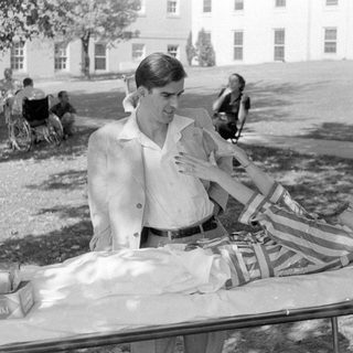Man standing beside a woman lying on a cot.