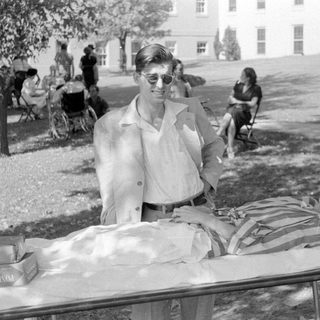 Man in suit standing by woman on a cot.