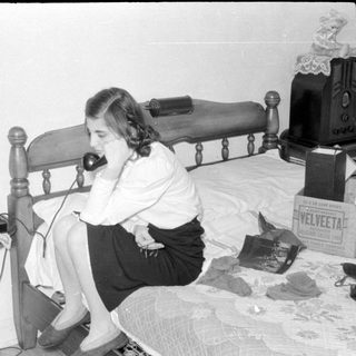 Woman on rotary phone sitting on a bed.