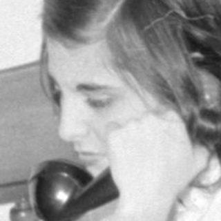 Woman on rotary phone sitting on a bed.