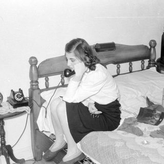 Woman on rotary phone sitting on a bed.