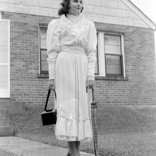 Woman in a ruffled dress standing by a house.