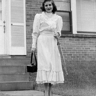 Woman in ruffled dress standing before a brick house.