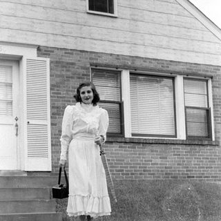 Woman in ruffled dress standing before a brick house.