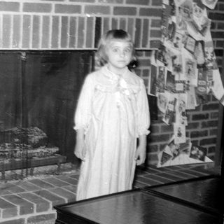 Girl in a dress standing by a brick fireplace.
