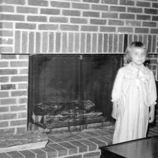 Girl in a dress standing by a brick fireplace.