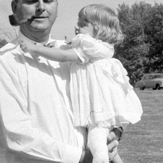 Man with pipe holding child in white dress.