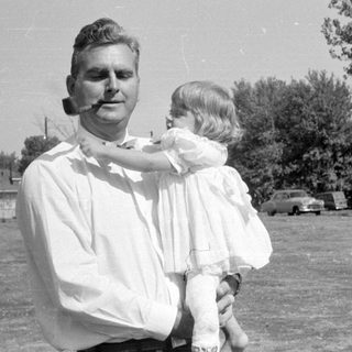 Man with pipe holding child in white dress.