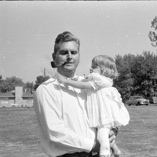 Man with pipe holding child in white dress.