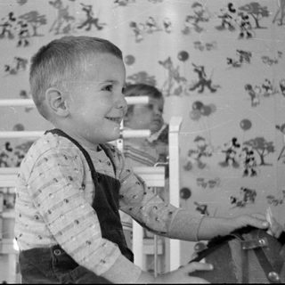 Young boy in overalls standing by a white crib.