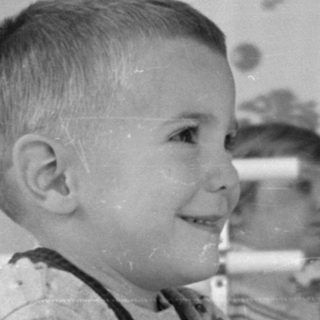 Young boy in overalls standing by a white crib.