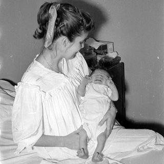 Woman in white dress holding an infant on a bed.