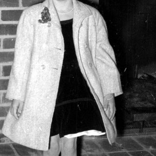 Girl in overcoat standing by a brick fireplace.