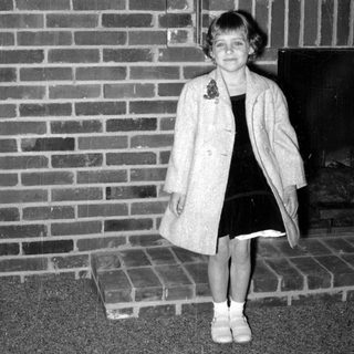 Girl in overcoat standing by a brick fireplace.